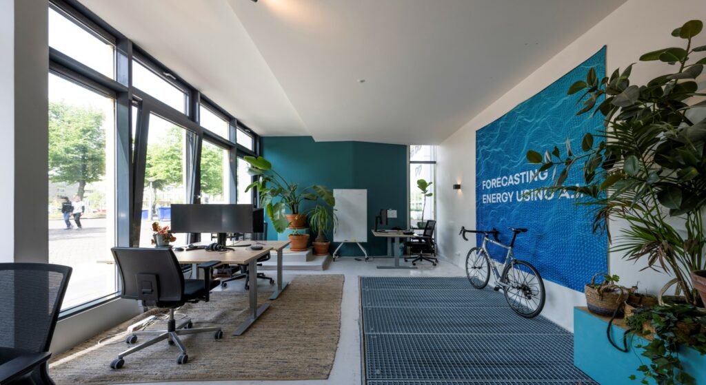 Moderne kantoorruimte aan de James Wattstraat met werkplekken, veel planten, een racefiets en een blauwe wand met de tekst "Forecasting Energy Using A.I.".